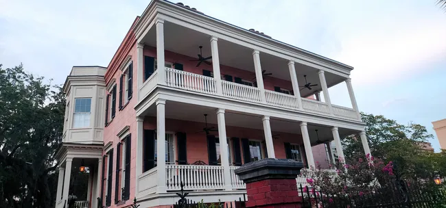 Historic pink house with multiple balconies and fans, surrounded by greenery in Savannah.