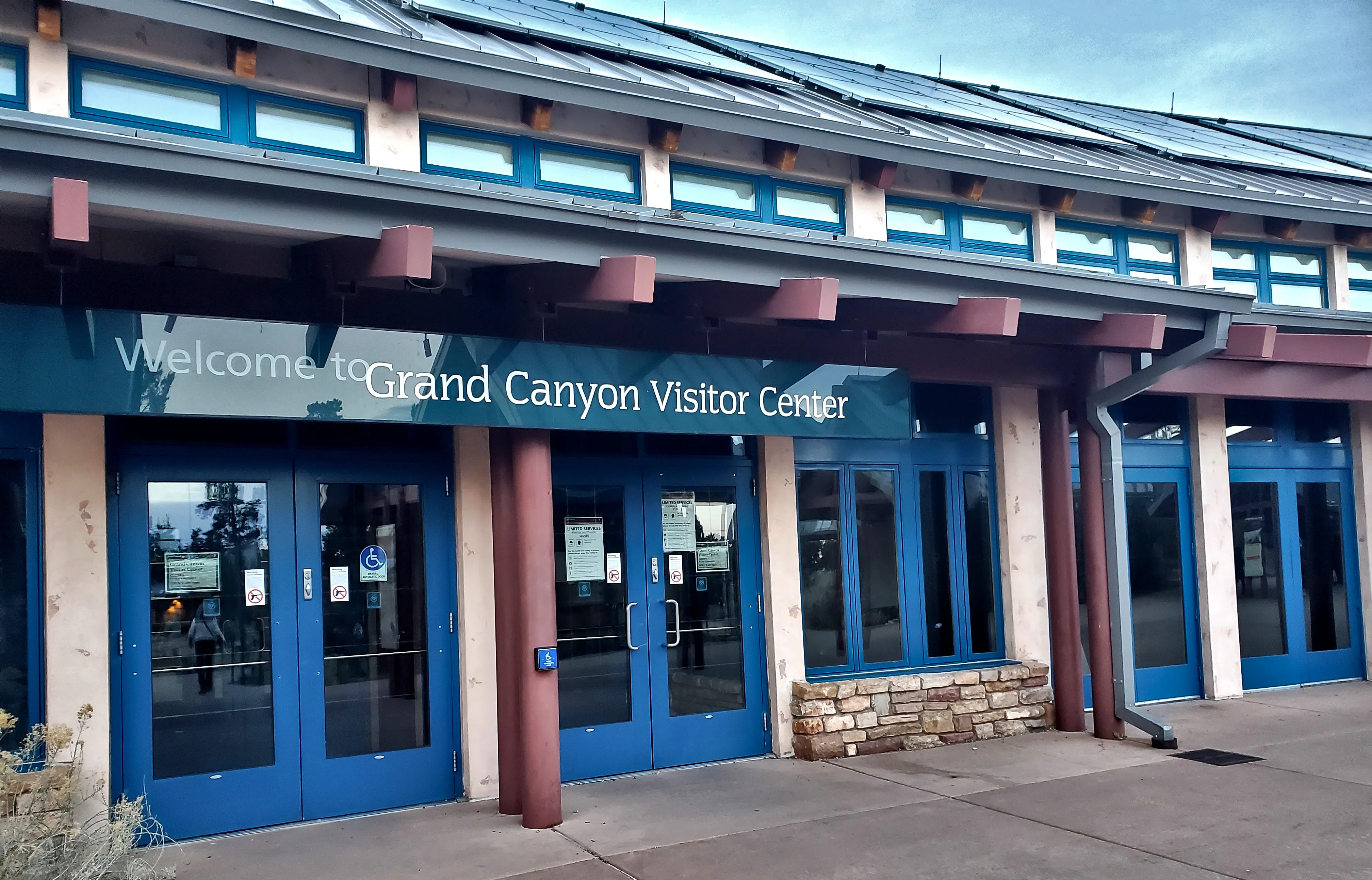 A modern visitor center entrance with blue double doors, large windows, and a sign above welcoming guests, set beneath an overhang supported by wooden beams.
