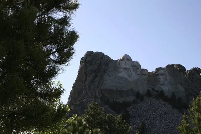 A view of Mount Rushmore National Memorial partially framed by pine trees, with clear detail of George Washington’s face and other U.S. presidents carved into the granite mountain.
