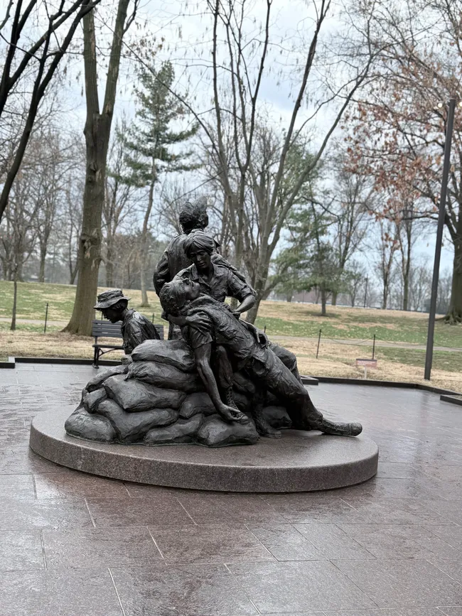 The Vietnam Women's Memorial, a bronze sculpture by Glenna Goodacre, located on the National Mall in Washington, D.C., near the Vietnam Veterans Memorial Wall. The statue depicts three military women — one cradling a wounded soldier across sandbags while another kneels nearby and a third looks skyward. The figures are rendered with expressive detail, conveying urgency and compassion. The sculpture sits on a round granite base on a wet, rain-slicked plaza. Bare winter trees and a park bench are visible in the overcast background.