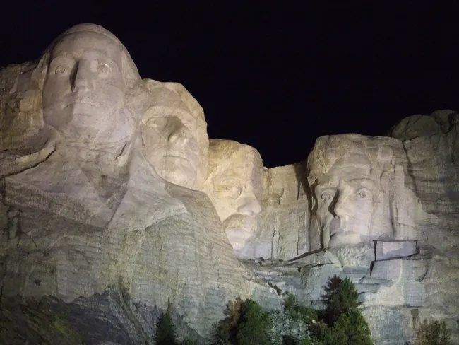 Nighttime view of Mount Rushmore National Memorial showing the illuminated stone carvings of four U.S. presidents.