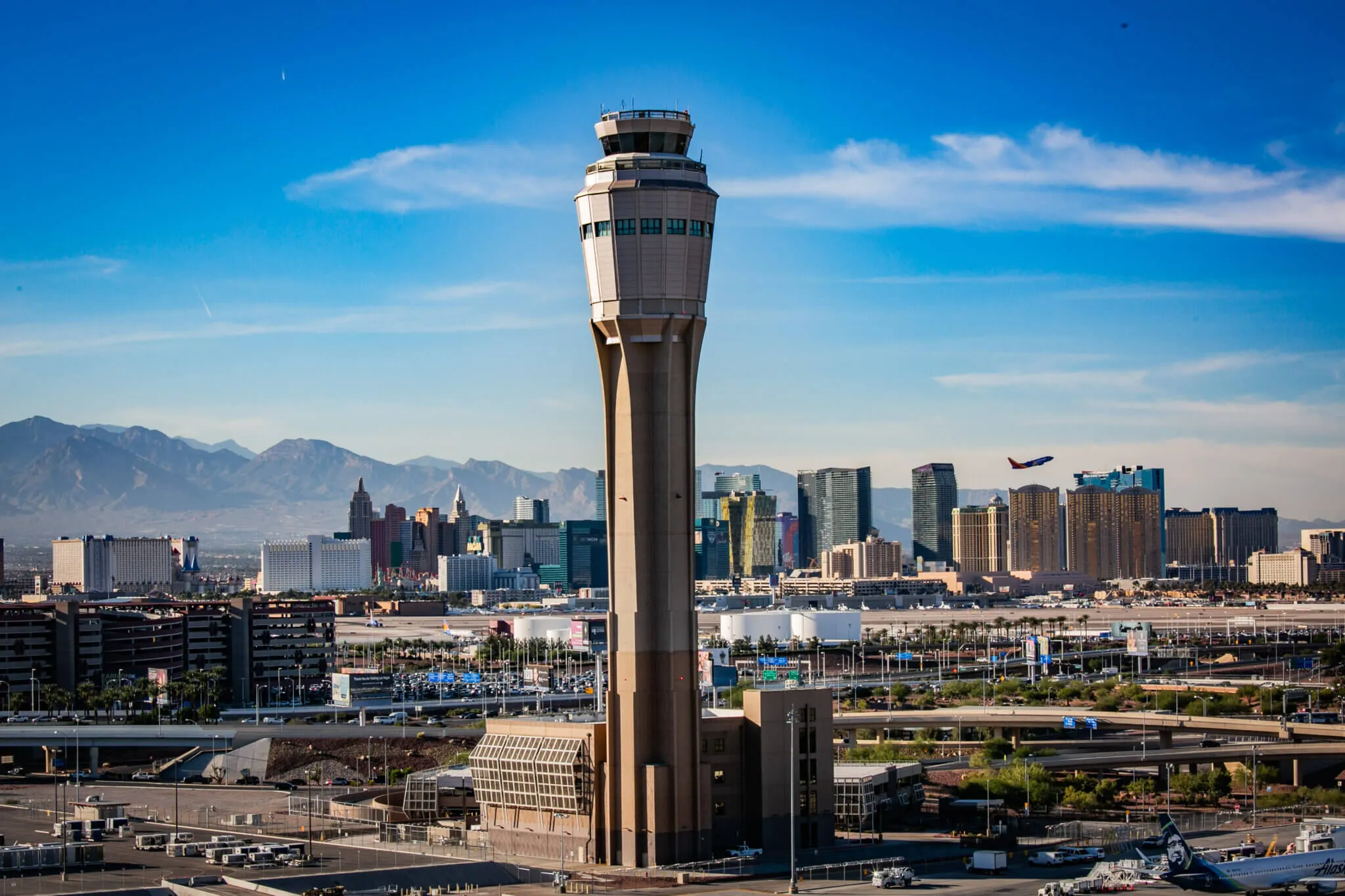 A high-angle view of a tall, tan-colored air traffic control tower standing in front of a dense skyline of colorful resort buildings and skyscrapers. In the background, hazy blue mountain peaks stretch across the horizon under a clear blue sky, with a commercial airplane seen taking off in the distance.
