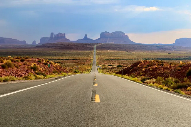 A straight highway stretches toward Monument Valley’s iconic buttes, framed by desert under a clear blue sky.