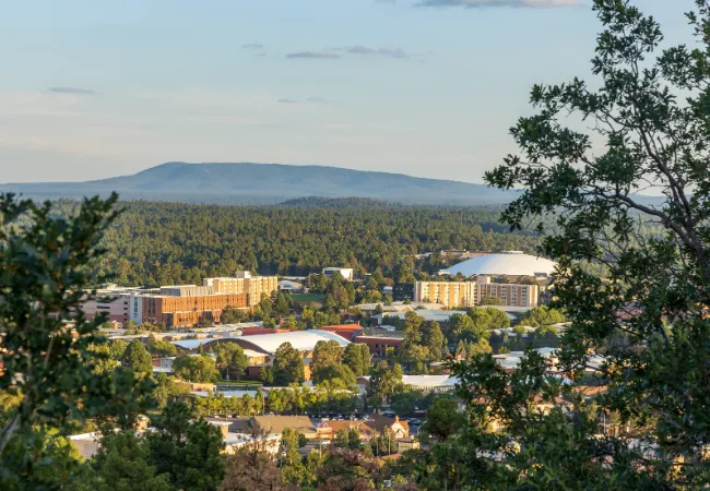An aerial view of the Northern Arizona University (NAU) campus in Flagstaff, featuring the Walkup Skydome nestled among dense pine forests with mountains in the background. The landscape showcases a mix of university buildings and lush greenery under a clear sky.