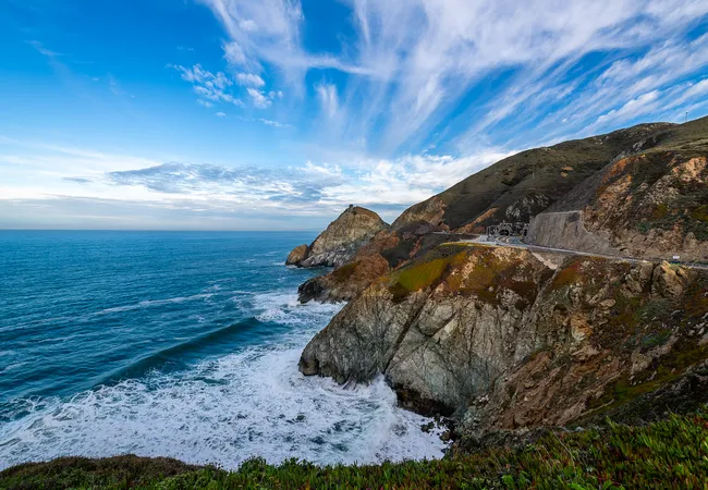 A winding coastal highway hugs steep ocean cliffs above crashing waves, with rugged headlands extending into the Pacific beneath a sky filled with dramatic wispy clouds.