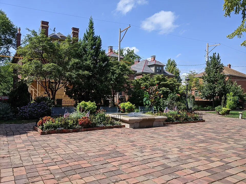 A sunny outdoor plaza in German Village, Columbus, Ohio, featuring a wide red brick paved courtyard. At the center is a low stone monument or plaque surrounded by raised garden beds enclosed by black iron fencing, filled with colorful flowers, shrubs, and small American flags. Blue park benches are visible among the greenery. In the background, historic red brick residential buildings with prominent chimneys are partially visible through a canopy of mature trees under a partly cloudy blue sky.