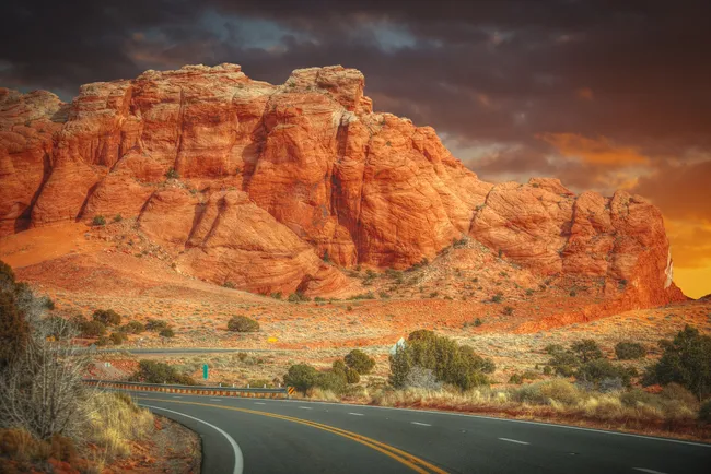 A winding highway curves toward towering red rock cliffs glowing under a dramatic sunset sky, with scattered desert shrubs in the foreground.