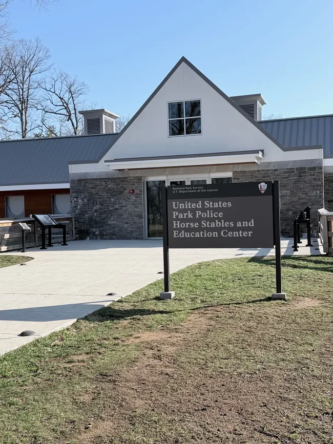 Exterior of the United States Park Police Horse Stables and Education Center, a modern building with a white gabled roofline, gray metal roof, and stone facade. A large black National Park Service sign in the foreground reads "National Park Service — U.S. Department of the Interior — United States Park Police Horse Stables and Education Center." Informational display panels are visible to the left along a concrete walkway. Bare trees and a clear blue sky are in the background.