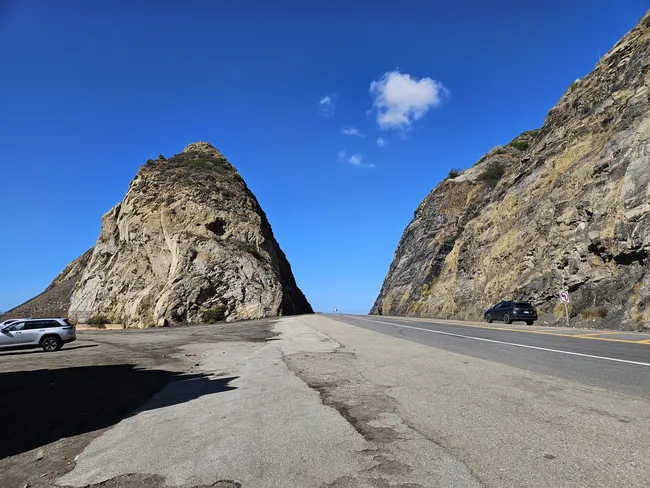 A wide-angle view of a paved highway passing between two massive, jagged rock formations. On the left, a large conical rock sits beside a small parking area with a silver SUV, while on the right, a steep rocky hillside rises directly from the road. The sky is a deep, clear blue with a single small white cloud floating above the gap in the rocks.