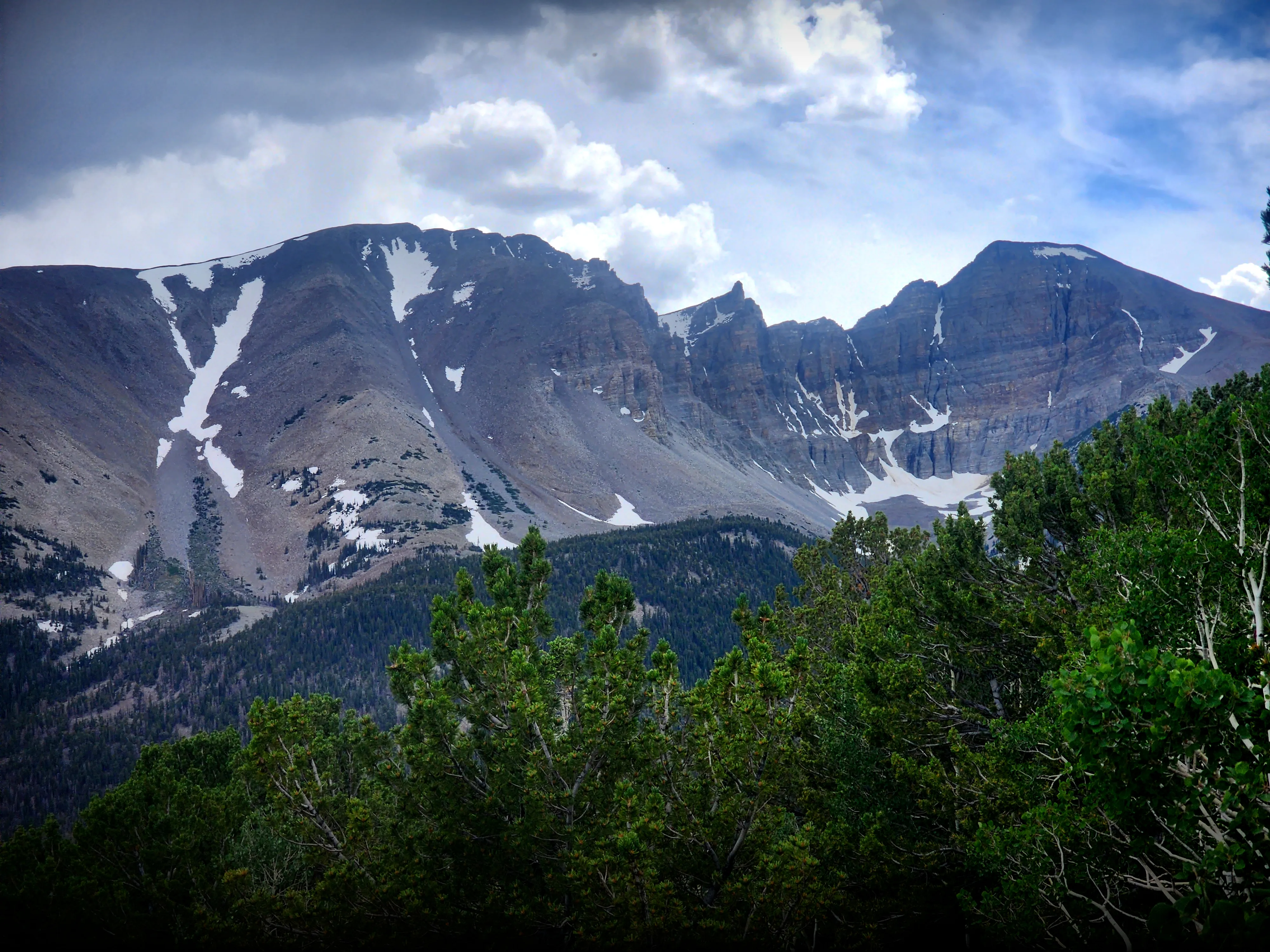 A dramatic mountain landscape view from the Wheeler Peak Overlook in Great Basin National Park, Nevada. The rugged, rocky peaks of Wheeler Peak rise steeply in the background, with patches of snow clinging to the upper slopes and gullies in midsummer. Dense subalpine conifer forests — including bristlecone pines — fill the foreground and middle slopes. Dramatic storm clouds build overhead against a partly blue sky.
