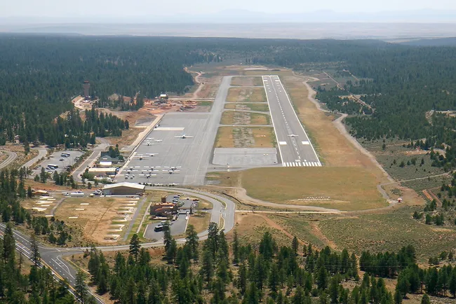 Small rural airport surrounded by forested terrain, featuring a single runway, taxiways, and a few parked aircraft.