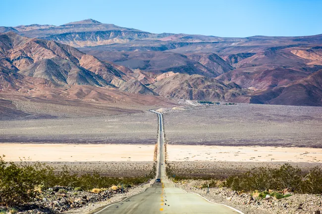 Long straight desert highway stretching toward rugged brown mountains under a clear blue sky.