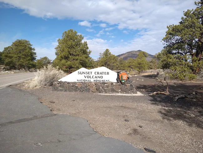 Entrance sign for Sunset Crater Volcano National Monument surrounded by pine trees and volcanic landscape.