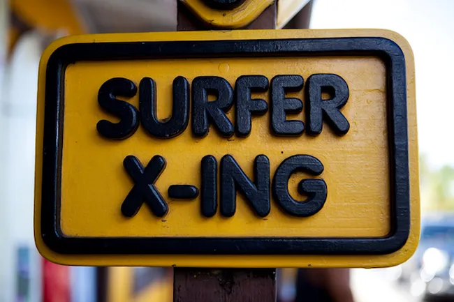 Yellow sign reading “Surfer X-ing” mounted on a post near a beach area.