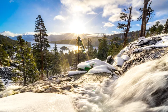 Snow-dusted waterfall flowing over rocks at sunrise with views of Emerald Bay, Lake Tahoe, and pine trees.