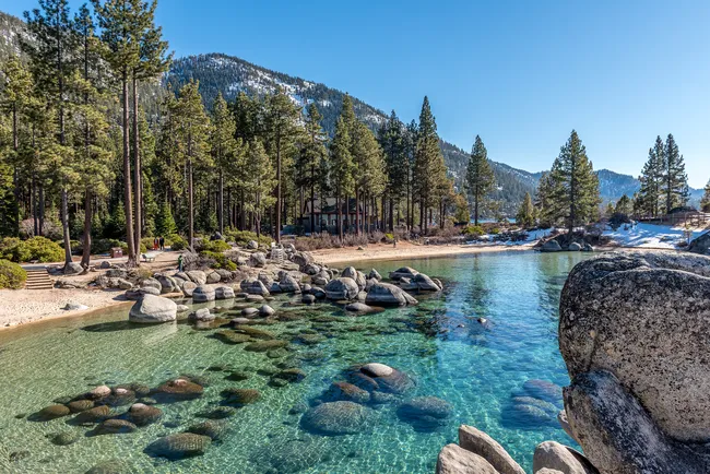 Clear turquoise water at Lake Tahoe’s Sand Harbor with smooth boulders, evergreen trees, and snow-covered mountains in the background.