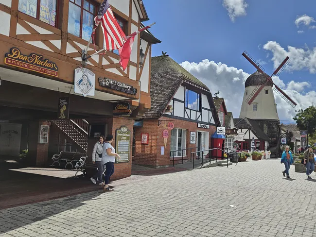 A wide-angle view of a village street paved with light grey cobblestones. On the left, a two-story building with white walls and brown timber framing features several shop signs and an American flag. In the background, a traditional white windmill with large brown blades stands prominently under a bright blue sky filled with large, fluffy white clouds.