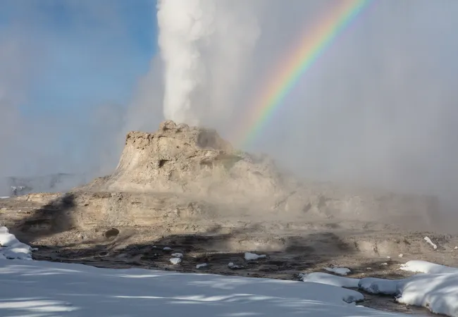 A steaming geyser rises from a rocky mound as a bright rainbow arcs through the mist, with patches of snow covering the ground.