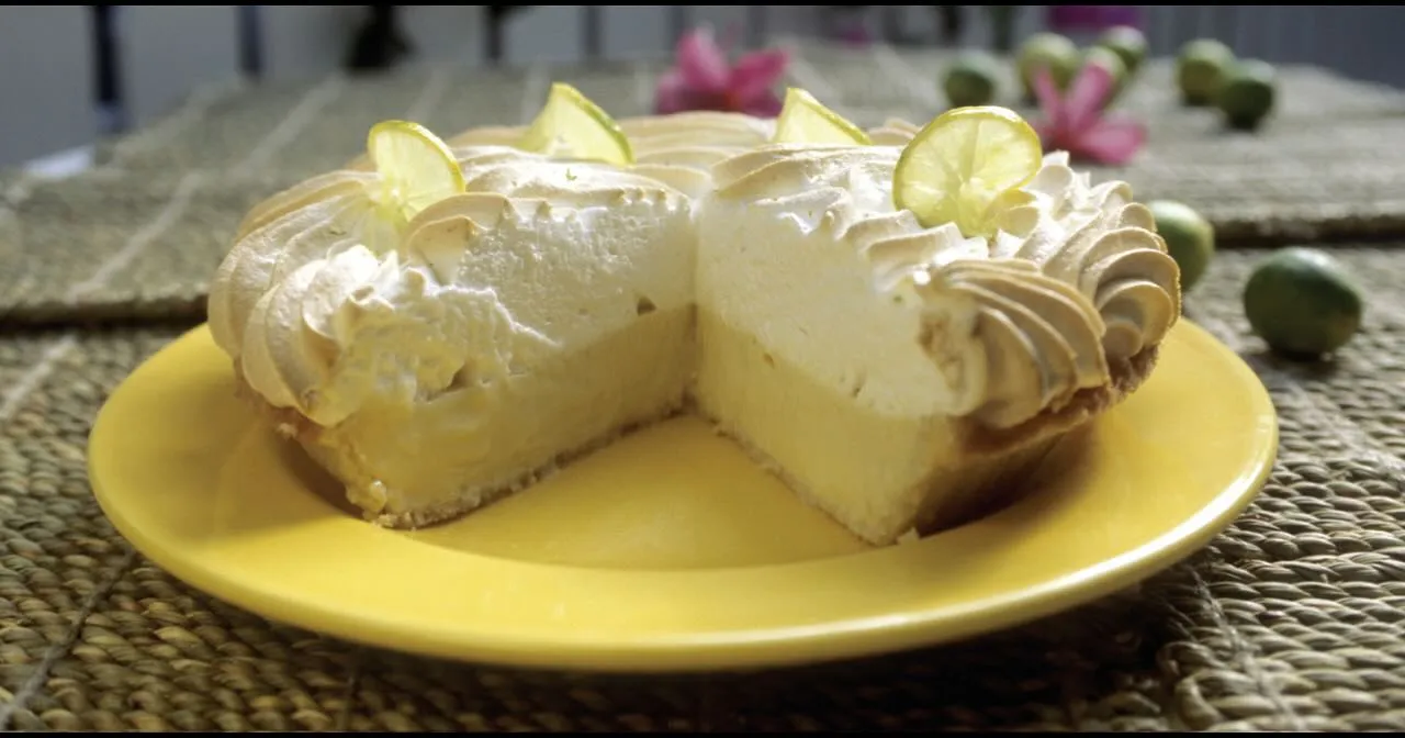 A eye-level, close-up shot of a key lime pie with a large slice removed to show the interior layers. The pie sits on a bright yellow plate atop a woven straw placemat. It features a thick, pale yellow citrus custard base and a massive layer of white meringue with lightly toasted ridges. Several thin, translucent lime wheels garnish the top. In the blurred background, small green key limes and pink flowers are scattered on the mat.