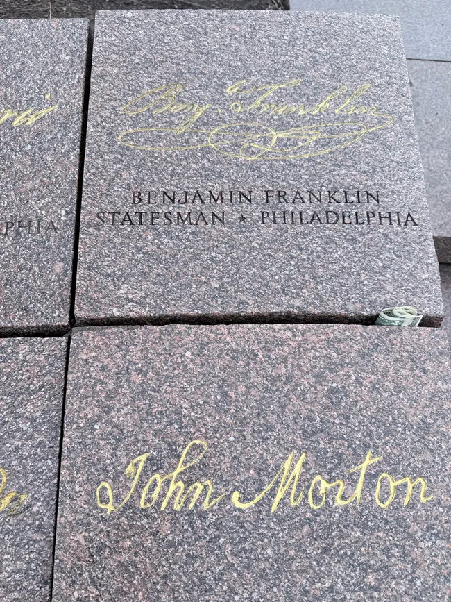 Close-up of polished granite memorial stones engraved with the signatures and names of signers of the Declaration of Independence. The central stone features the gold cursive facsimile signature of Benjamin Franklin, with the engraved text "Benjamin Franklin — Statesman · Philadelphia" below it. A partially visible stone to the lower right bears the gold cursive signature of John Morton. A folded dollar bill has been tucked into the seam between the stones as a tribute. The stones are part of a commemorative monument, likely in Washington, D.C.
