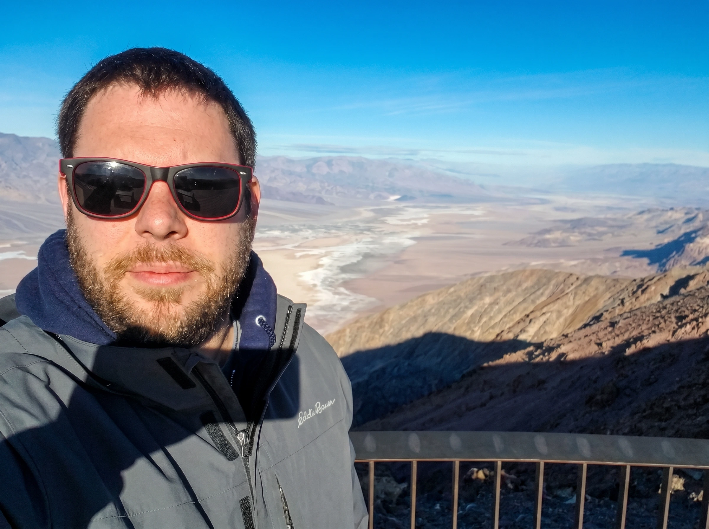 A video screenshot showing a man with short dark hair and a beard, wearing dark sunglasses with red frames and a gray Eddie Bauer jacket over a blue hoodie, taking a selfie at a high-elevation overlook. Behind him stretches a vast desert valley with a dry salt flat visible far below, surrounded by rugged brown mountain ridges — consistent with the view from Dante's View in Death Valley National Park. A clear blue sky fills the upper portion of the frame. Video playback controls are visible at the bottom of the screen.