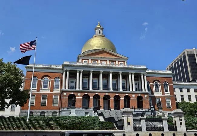 The Massachusetts State House features its golden dome, red brick facade, and grand staircase under a sunny blue sky.