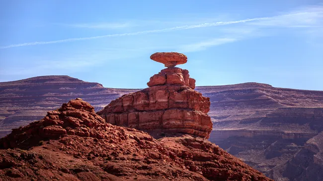 A rock formation shaped like an upside-down sombrero balances atop layered red cliffs in Utah.