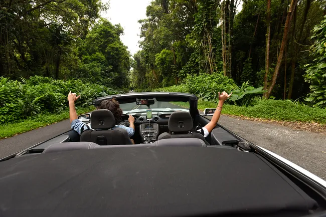 Two people driving through a lush tropical forest in a black convertible with the top down, both raising their hands in a shaka sign.
