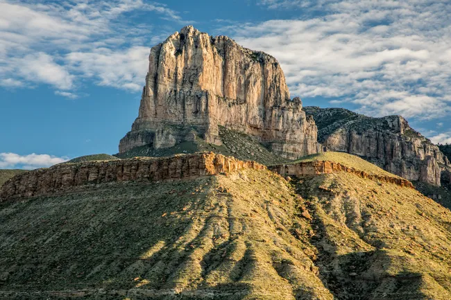 Alt text: Massive limestone cliffs of El Capitan rise above layered desert ridges and rugged foothills, illuminated by warm sunlight beneath a partly cloudy sky.