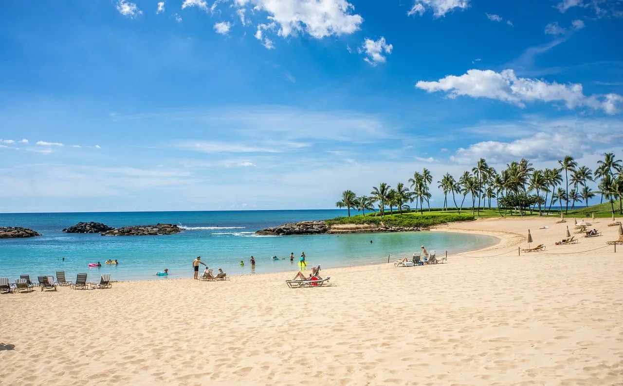 People relaxing and swimming at a calm, crescent-shaped beach lagoon with golden sand and palm trees.