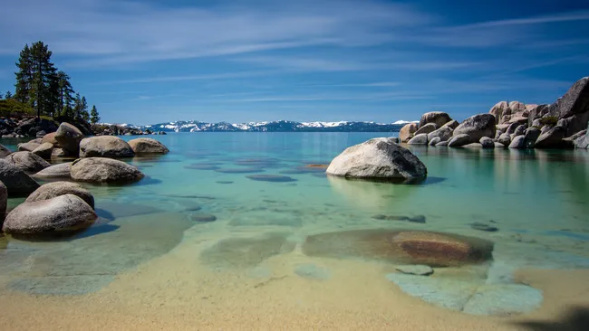Clear turquoise lake with smooth boulders, sandy shallows, and snow-capped mountains in the distance under a bright sky.