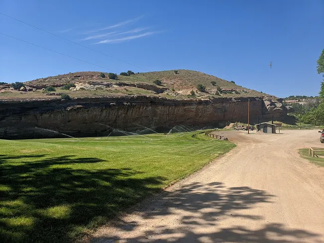 Dirt road and grassy area at Medicine Lodge Archaeological Site in Wyoming, with a dramatic sandstone cliff in the background and sprinklers watering the lawn on a sunny day.