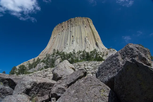Towering view of Devils Tower National Monument in Wyoming, a striking geological formation with vertical columns rising above a field of large boulders under a clear blue sky.