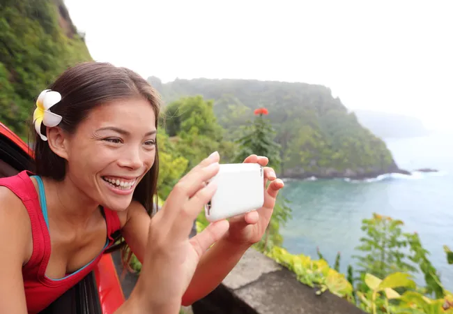 Smiling woman with a plumeria flower in her hair leans from a car window to take a scenic coastal photo.