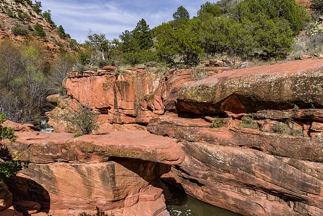 Layers of red sandstone cliffs and a small canyon pool surrounded by desert vegetation under a bright sky.