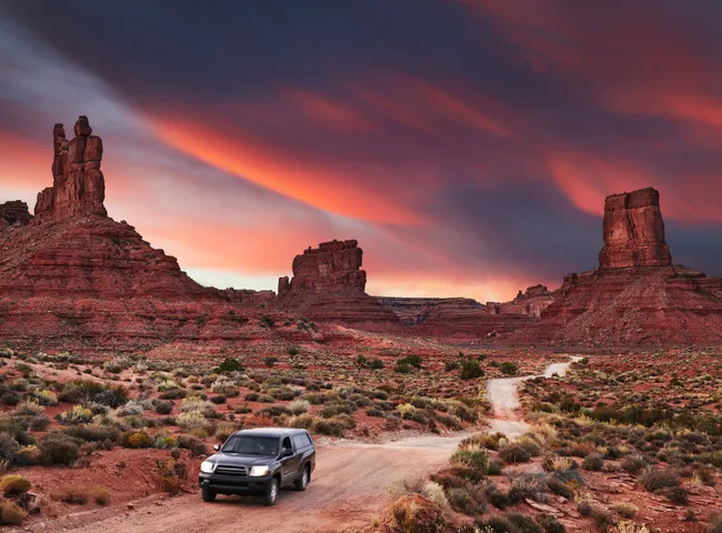 SUV driving on a dirt road through red rock formations in Valley of the Gods at sunset.