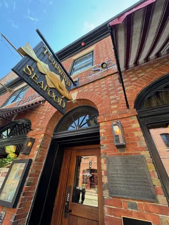 Front view of Turner’s Seafood restaurant in Salem, MA, featuring a golden fish sign hanging above a brick archway and a striped awning overhead.