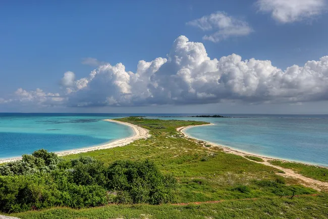 A scenic view of Bush Key at Dry Tortugas National Park with a sandy shoreline, lush greenery, and turquoise waters under a partly cloudy sky.