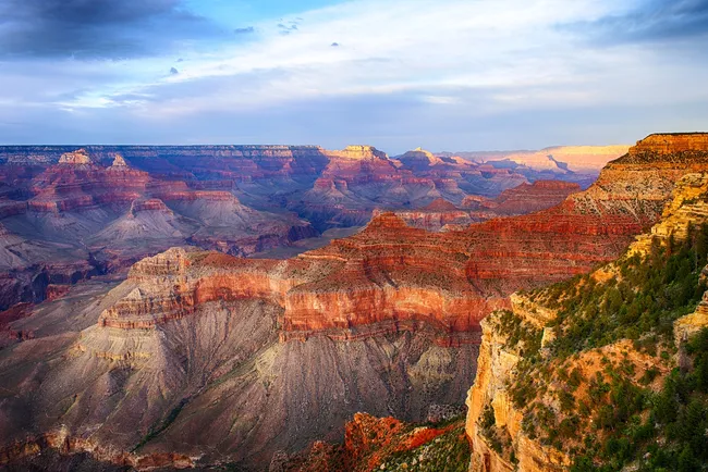 Layered red and orange canyon walls stretch across a vast landscape under a soft, cloudy sky, with sunlight highlighting ridges and cliffs.