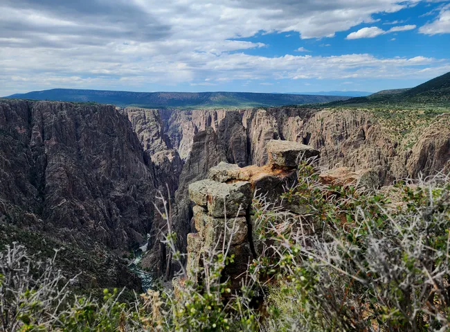 Dramatic view into Black Canyon of the Gunnison with jagged rock formations, steep canyon walls, and the Gunnison River flowing far below under a partly cloudy sky.
