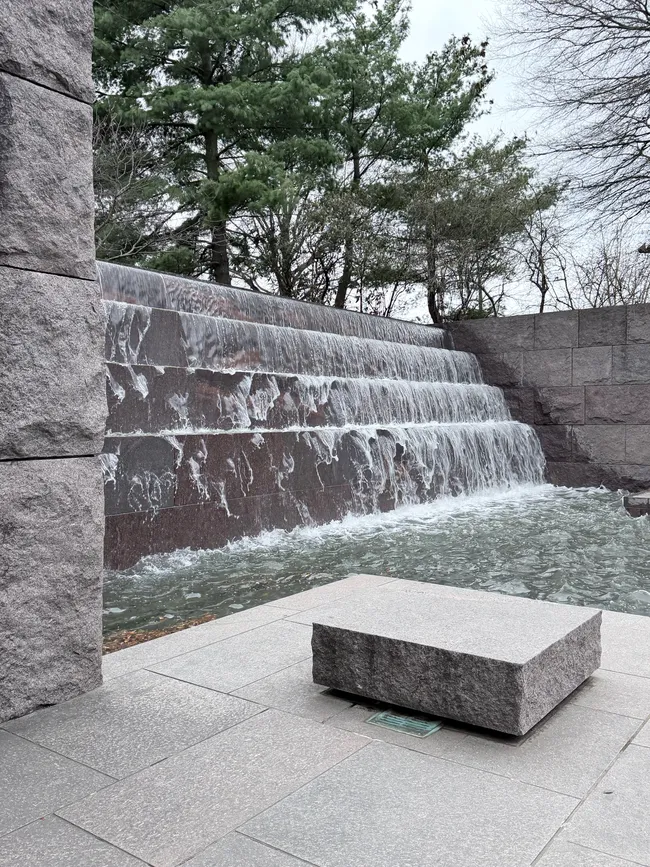 A cascading waterfall flowing over a wide, multi-tiered rough-textured red granite wall into a shallow reflecting pool at the Franklin Delano Roosevelt Memorial in Washington, D.C. A small square granite block sits in the foreground on a smooth stone plaza. Large granite wall panels flank the sides, and a mix of evergreen and bare deciduous trees rise above the memorial walls under an overcast gray sky.