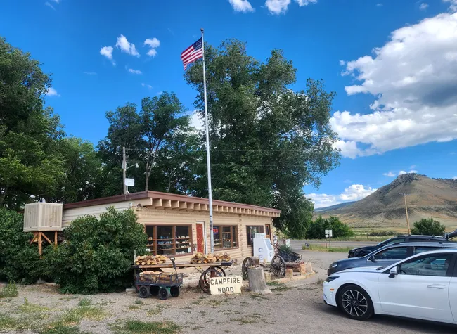 A small general store near Black Canyon of the Gunnison with an American flag flying above, stacks of campfire wood out front, and a scenic mountain landscape in the background.