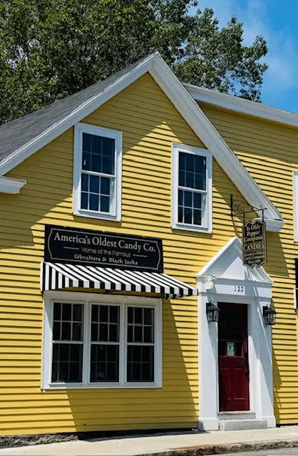 Yellow clapboard building with a red door and striped awning, home to America’s Oldest Candy Company.