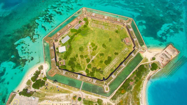 Alt text: Aerial view of Fort Jefferson, a large hexagonal brick fortress surrounded by a moat and bright turquoise waters, situated on a small island connected by a narrow causeway.
