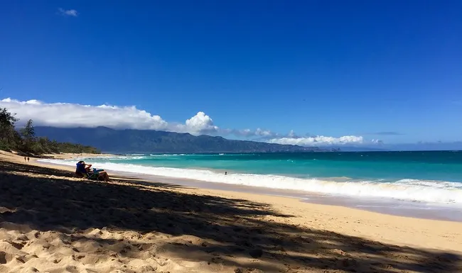 Sunny beach with turquoise waves, golden sand, and distant mountains under a clear blue sky.
