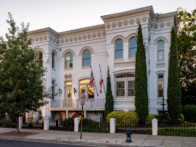 Exterior of Florida House, a stately white two-story building in Washington, D.C., serving as Florida's official state hospitality house. The Italianate-style structure features arched windows, decorative cornice detailing, and a small front balcony where an American flag and the Florida state flag are displayed. Tall slender cypress trees flank the building, and a black iron fence lines the front. The warm glow of interior lighting is visible through the windows at dusk.