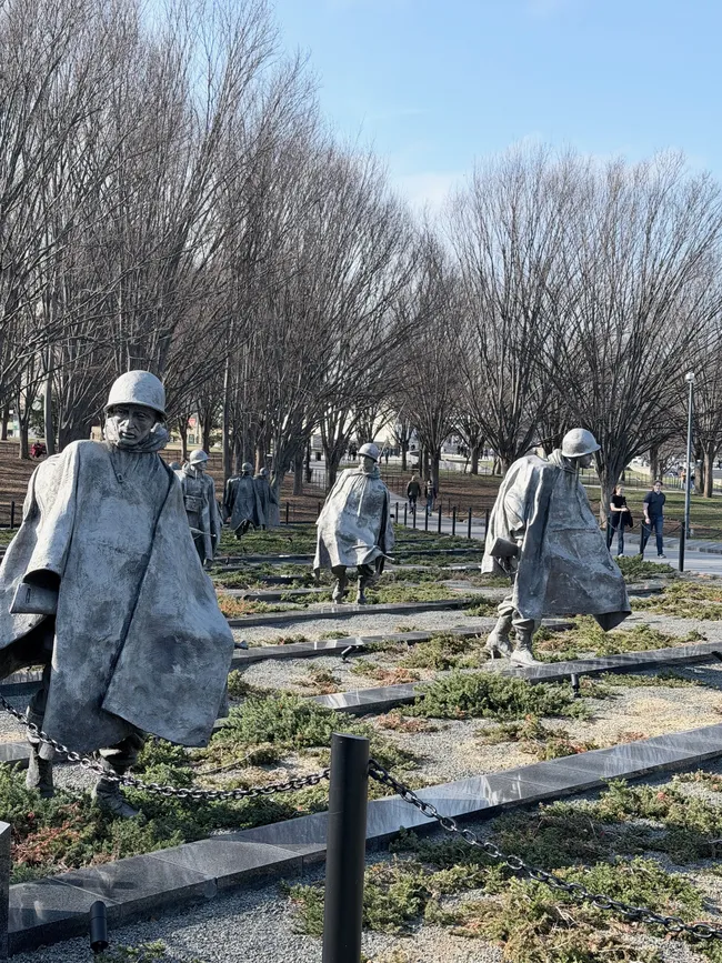 The Korean War Veterans Memorial on the National Mall in Washington, D.C., featuring a column of stainless steel soldier statues advancing through a field of low juniper ground cover interspersed with granite strips. The larger-than-life figures are dressed in full combat gear — helmets, ponchos, and weapons — with weathered, determined expressions. A chain barrier lines the perimeter. Bare winter trees form a dense backdrop, and several visitors are visible in the distance on a clear, sunny day.
