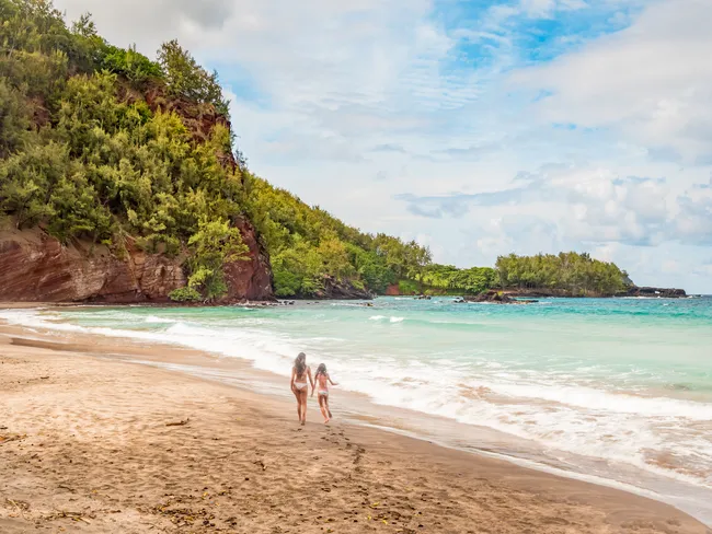 Two people walking along Koki Beach in Maui, with red cliffs, turquoise water, and lush greenery.
