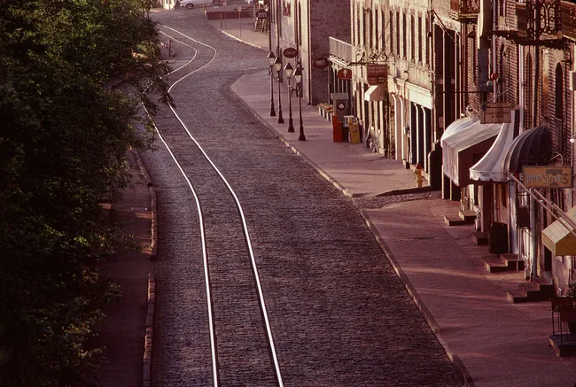 Cobblestone street with railway tracks, flanked by buildings and street lamps, likely near The Shrimp Factory.