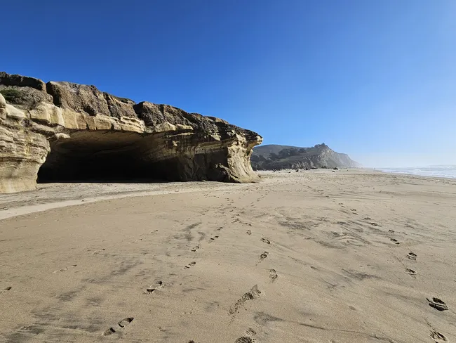 A broad sandy beach stretches beneath sculpted coastal bluffs, featuring a large sea cave carved into the cliff face. Footprints trail across the sand toward the shoreline, while rugged headlands and the ocean extend into the distance under a clear blue sky.